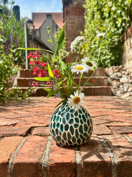 Vase mit Blumen auf einer Ziegelsteinterrasse in einem Garten.